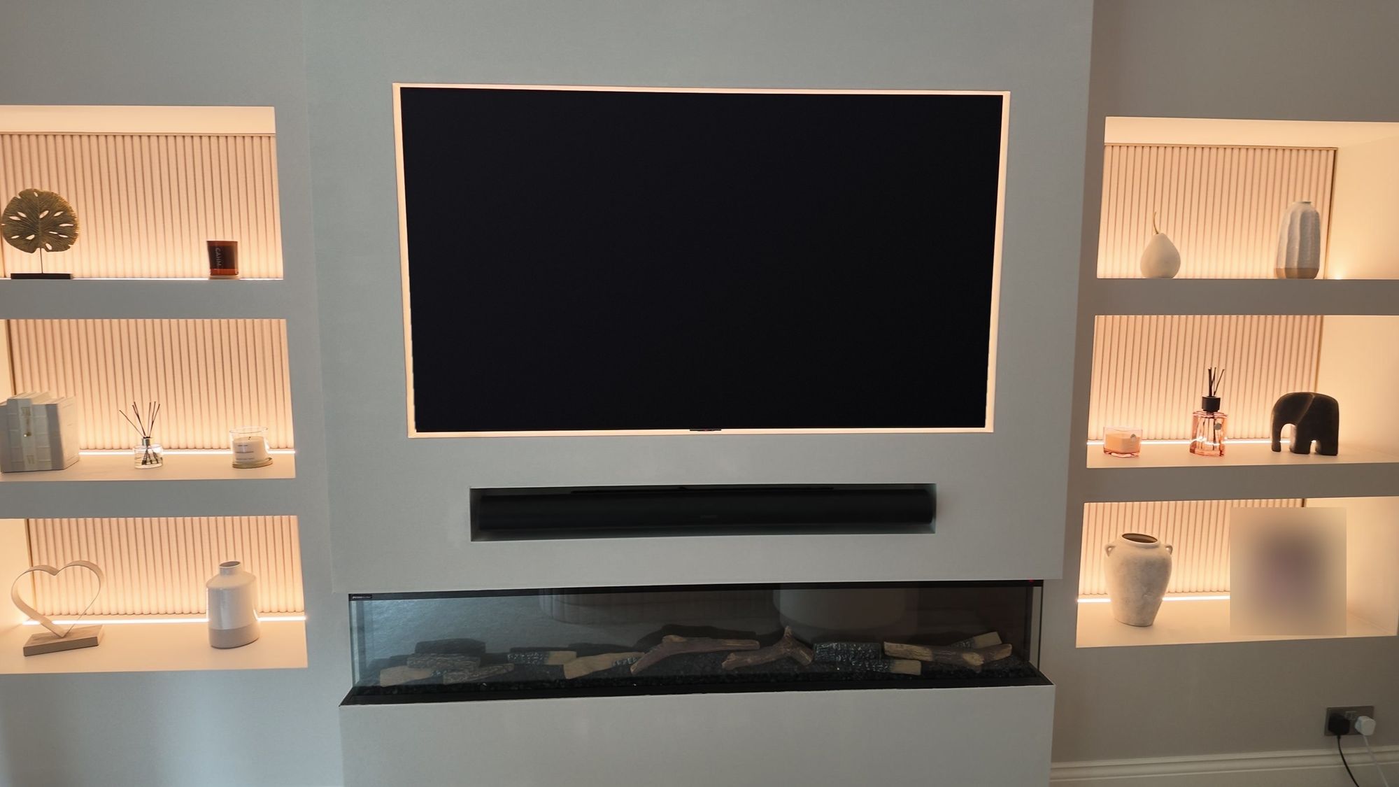 Finished media wall: TV in a backlit recess, built-in soundbar, long electric fire, lit shelf niches either side
